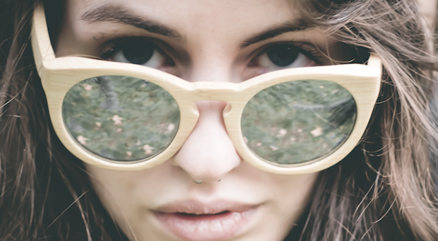 Primer plano de una persona de piel clara que lleva unas grandes gafas de sol redondas de madera. Los cristales reflejan una escena exterior verde y frondosa. La persona tiene los labios ligeramente separados y el pelo castaño le cubre parcialmente la cara.