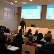 Un home està dret parlant per un micròfon davant d'un públic en una sala de conferències. Les diapositives de la presentació sobre els avenços en el tractament de l'ull sec es projecten en pantalles darrere seu. Diverses persones escolten atentament.