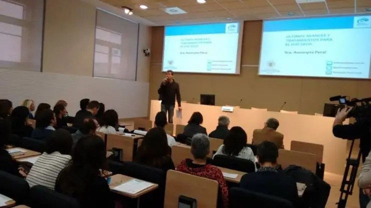 Un home està dret parlant per un micròfon davant d'un públic en una sala de conferències. Les diapositives de la presentació sobre els avenços en el tractament de l'ull sec es projecten en pantalles darrere seu. Diverses persones escolten atentament.