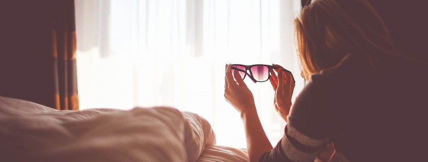 Una persona amb pijama estirada en un llit amb unes ulleres de sol apuntant cap a una finestra il·luminada pel sol, amb una llum suau que entra a través de les cortines transparents; l'ambient acollidor de l'habitació insinúa una visió nítida, com si s'aconseguís amb Orto-K o llenties.