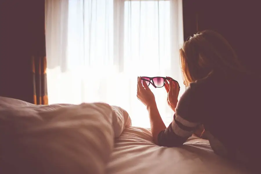 Una persona amb pijama estirada en un llit amb unes ulleres de sol apuntant cap a una finestra il·luminada pel sol, amb una llum suau que entra a través de les cortines transparents; l'ambient acollidor de l'habitació insinúa una visió nítida, com si s'aconseguís amb Orto-K o llenties.
