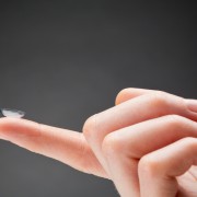 Primer pla d'una mà que sosté una lent transparent Orto-K a la punta del dit índex sobre un fons llis i fosc.