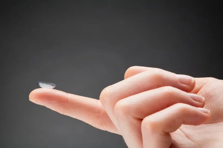 Primer pla d'una mà que sosté una lent transparent Orto-K a la punta del dit índex sobre un fons llis i fosc.