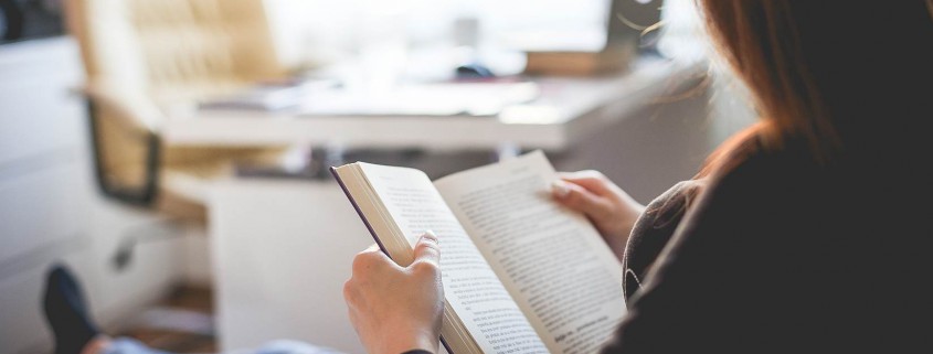 Una persona amb els cabells llargs seu a l'interior, relaxant-se i llegint un llibre com a part de la seva imatge visual de teràpia. El fons està suaument enfocat, mostrant un escriptori, una cadira i una finestra, creant una atmosfera tranquil·la i acollidora.