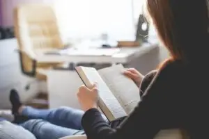 Una persona amb els cabells llargs seu a l'interior, relaxant-se i llegint un llibre com a part de la seva imatge visual de teràpia. El fons està suaument enfocat, mostrant un escriptori, una cadira i una finestra, creant una atmosfera tranquil·la i acollidora.