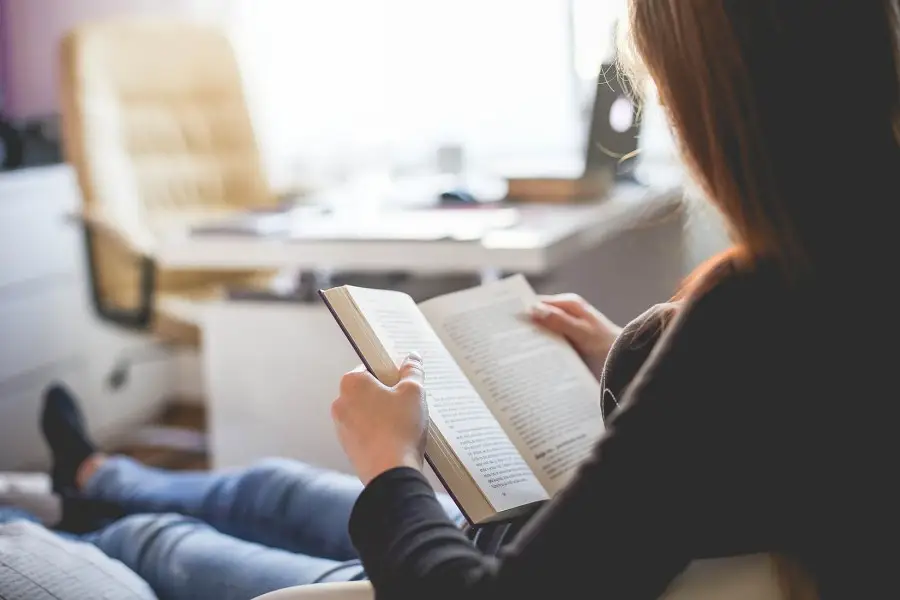 Una persona amb els cabells llargs seu a l'interior, relaxant-se i llegint un llibre com a part de la seva imatge visual de teràpia. El fons està suaument enfocat, mostrant un escriptori, una cadira i una finestra, creant una atmosfera tranquil·la i acollidora.