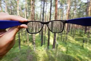 Una mà sosté unes ulleres davant d'un bosc, fent que els arbres apareguin nítids i clars a través de les lents mentre que la zona circumdant roman borrosa, il·lustrant vívidament els efectes de la miopia.