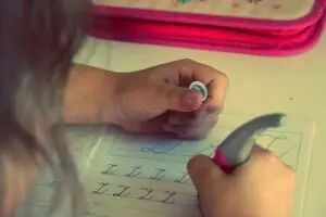 Un nen practica l'escriptura cursiva amb un bolígraf en un quadern com a part de Teràpia Visual. Només es veuen les mans del nen i una part del quadern sobre una superfície blanca, amb una caixa rosa al fons.